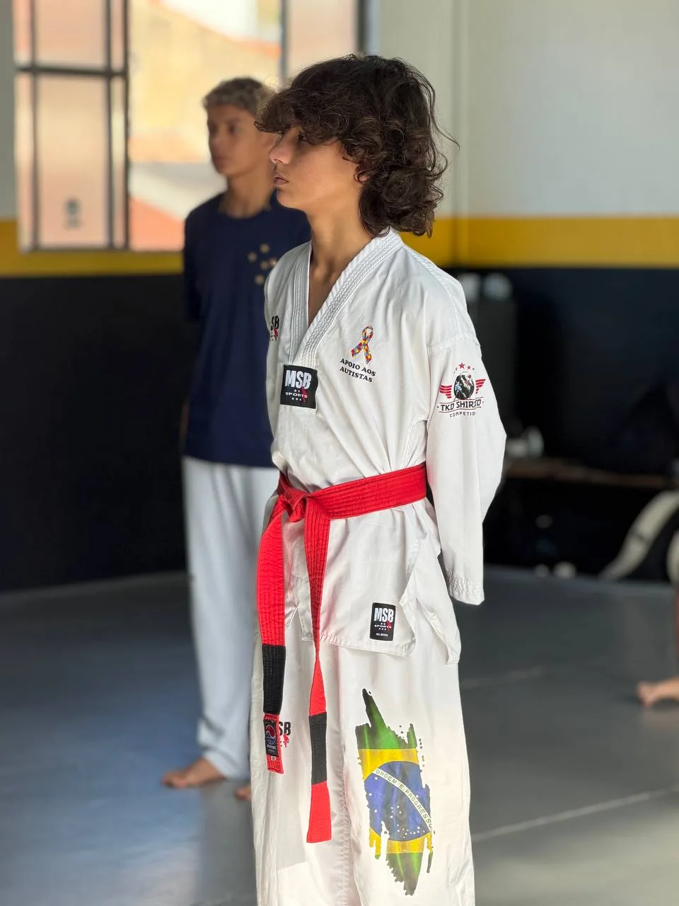 Atleta faixa vermelha do Instituto Shirato perfilado no dojang, de dobok e com a bandeira do Brasil na calça